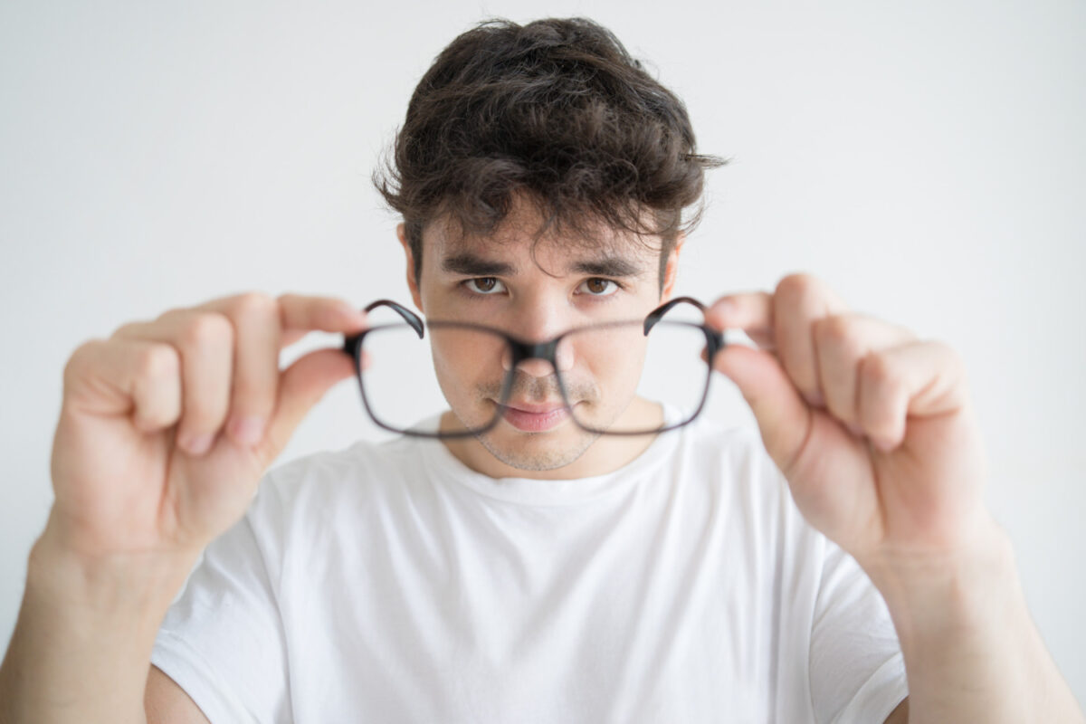 Hombre joven observando sus gafas con dificultad para enfocar, gesto asociado a visión borrosa o posibles problemas visuales.